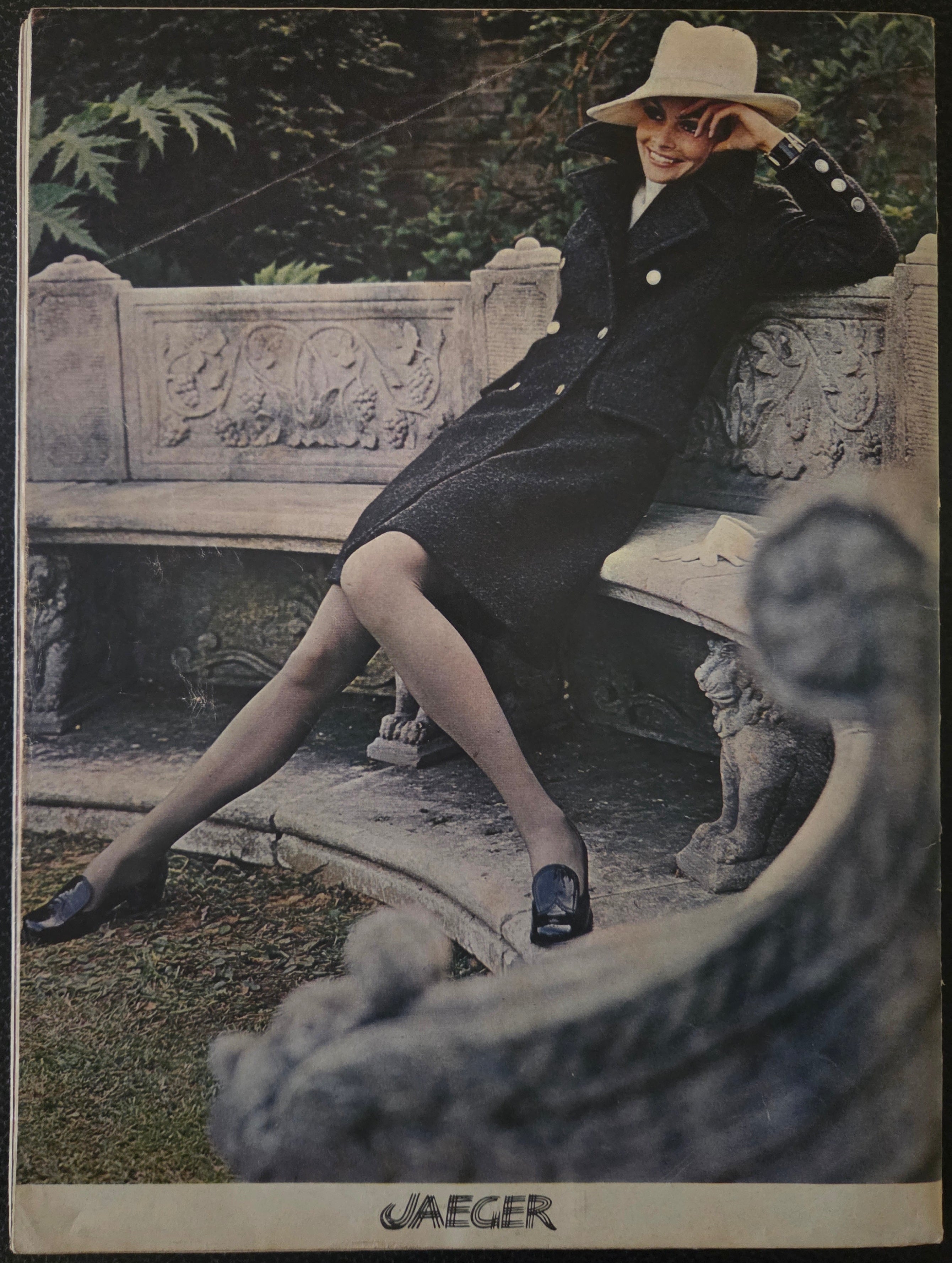 Vintage Jaeger advert with model on stone bench in black coat and white hat.
